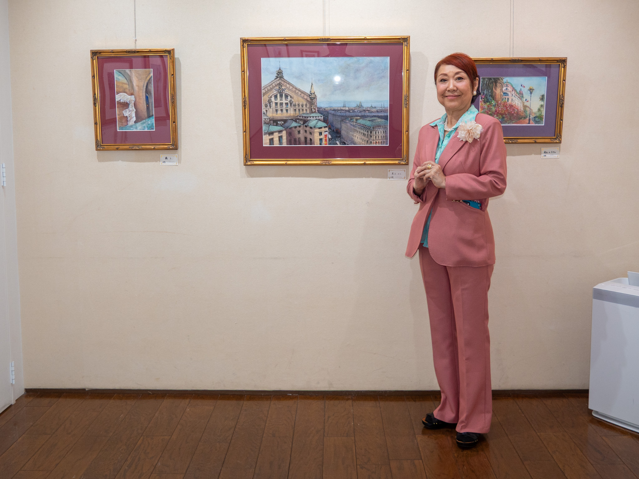 個展会場で展示作品と並ぶ小原けい子。ピンクの服を着て、穏やかに微笑んでいる